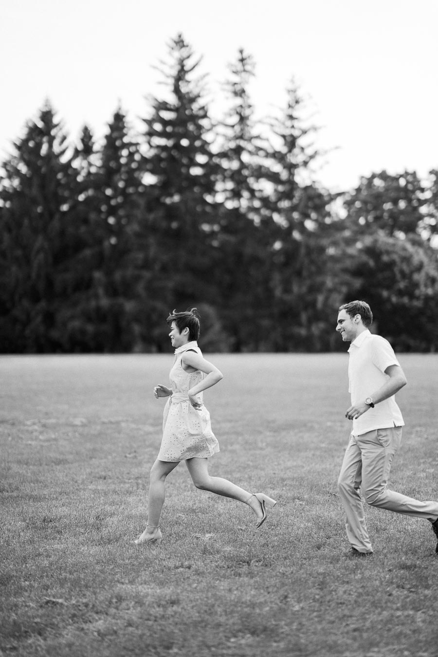 Black and white photo of a couple running playfully through an open field with tall trees in the background, symbolizing joy and freedom in nature.