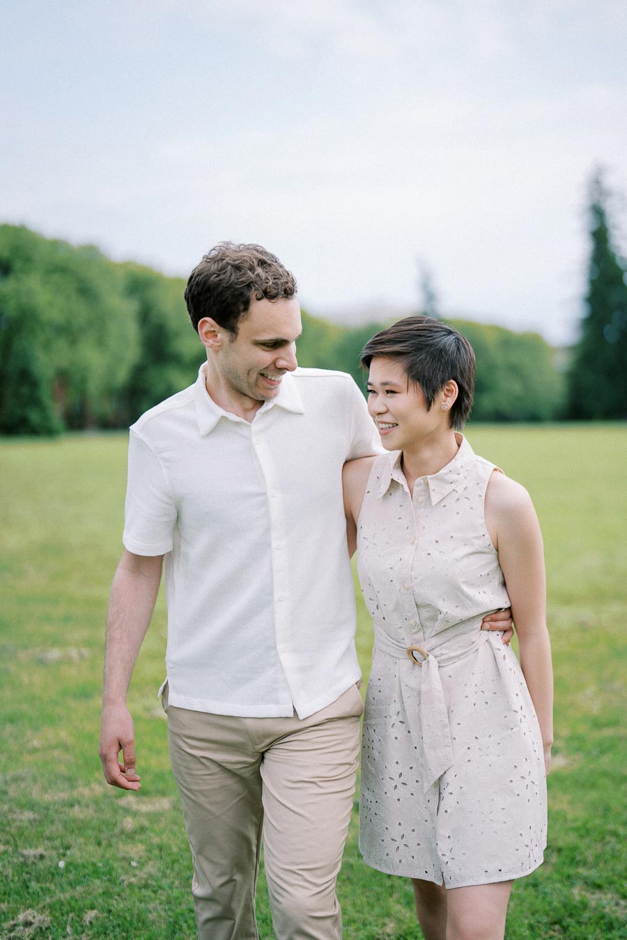 A happy couple walking arm-in-arm in a lush green park on a sunny day, both wearing light-colored summer outfits and smiling at each other.