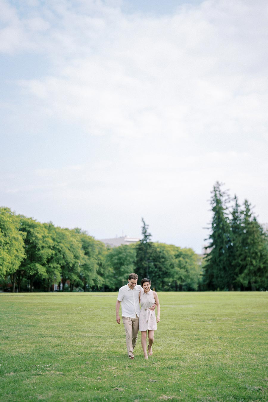 A couple walking arm in arm in a lush green park under a cloudy blue sky.