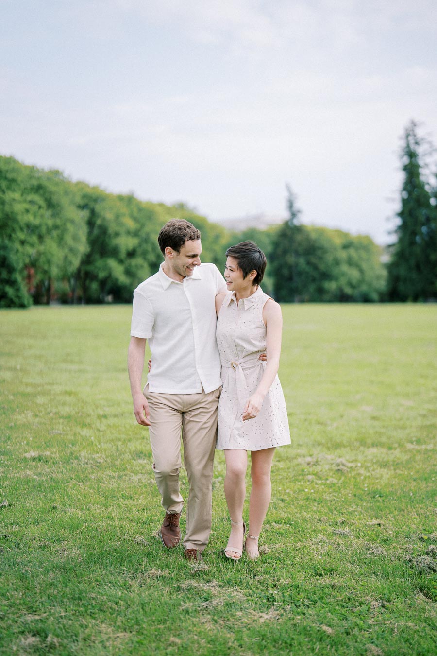 A couple walking together in a lush green park, smiling at each other against a backdrop of trees and a clear sky.