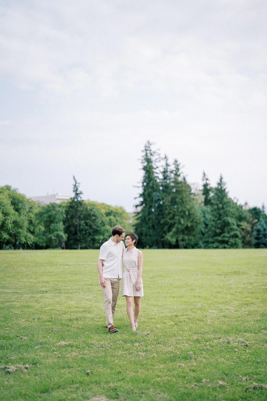 A couple strolling across a lush green park with tall trees in the background, under a partly cloudy sky.