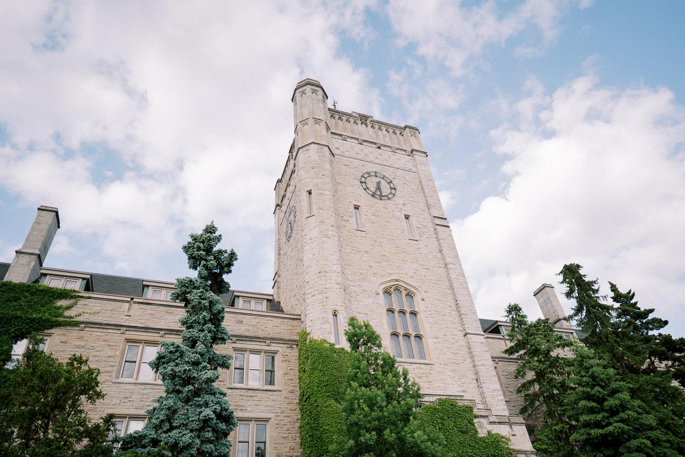 Historic stone clock tower with ivy-covered walls against a partly cloudy sky, flanked by tall green trees.