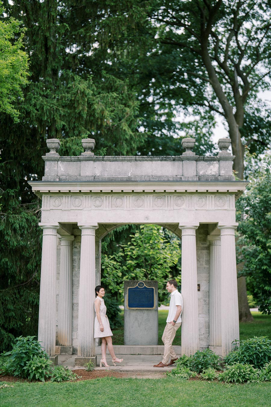 A couple stands smiling under a historic stone gazebo surrounded by lush greenery in a scenic park setting.