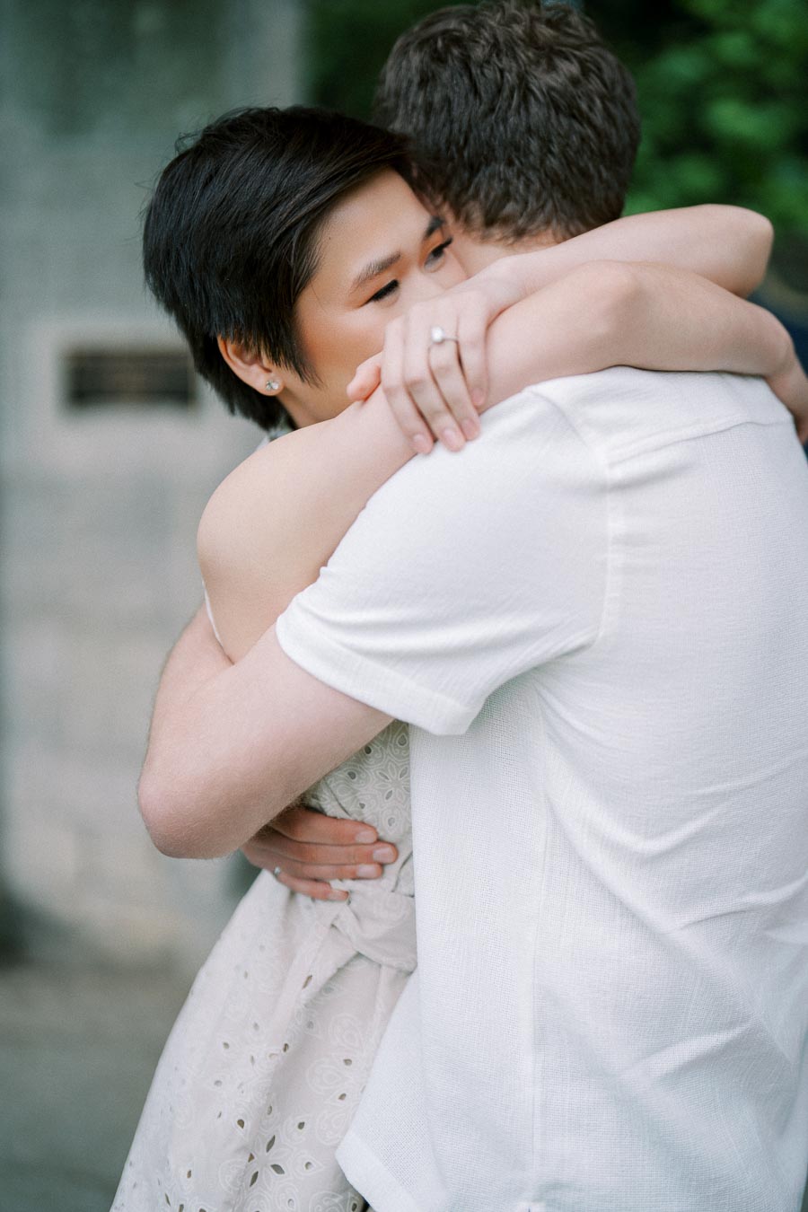 A couple embracing with visible engagement ring, capturing a romantic moment in a soft-focus outdoor setting.