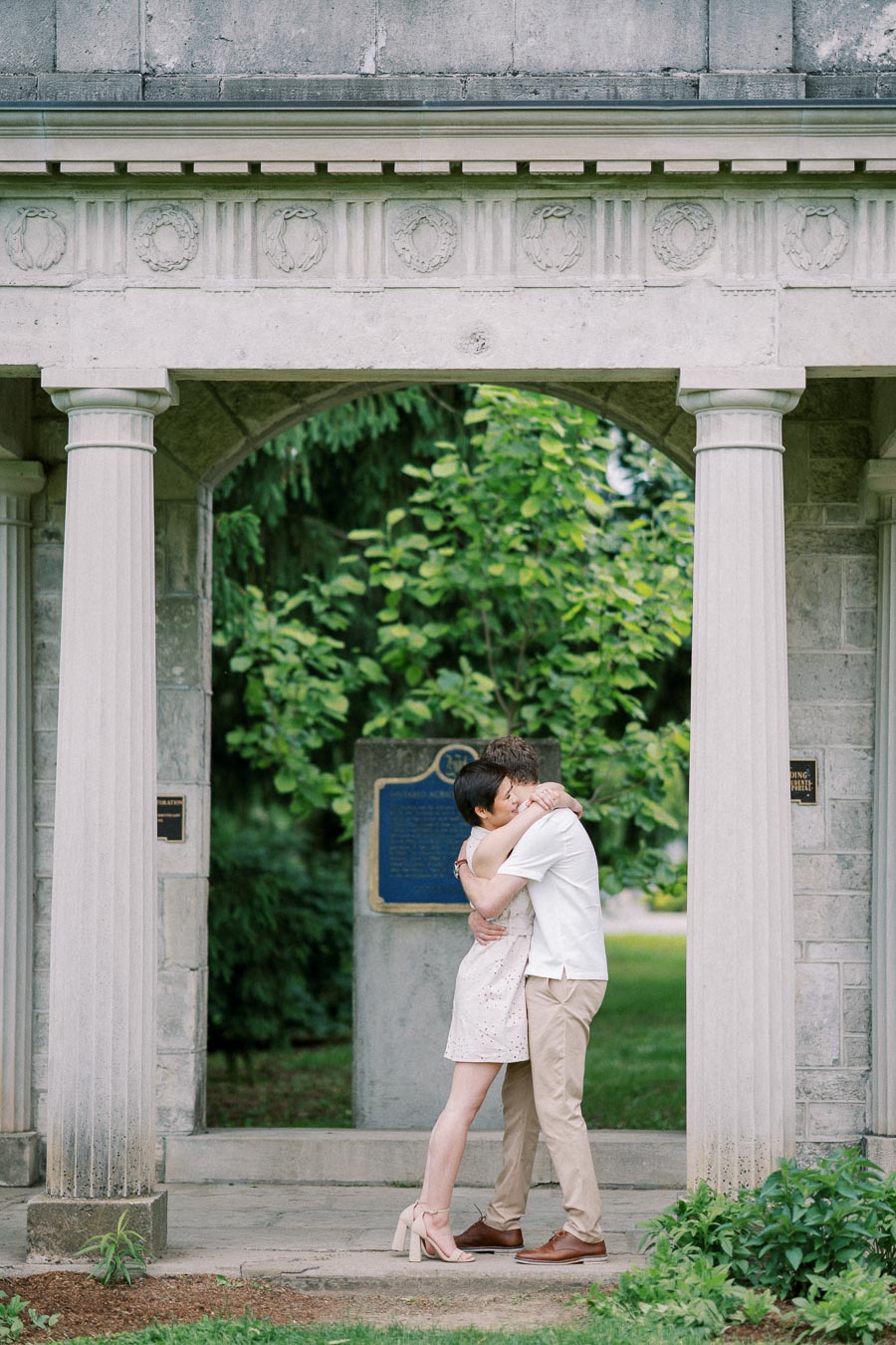 A couple sharing an intimate embrace under a stone archway, surrounded by lush greenery and historical architecture.