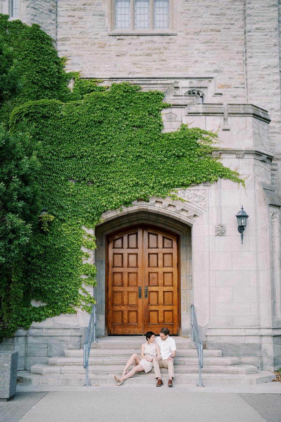 A couple sitting on stone steps in front of a historical building with a large wooden door, partially covered in green ivy.