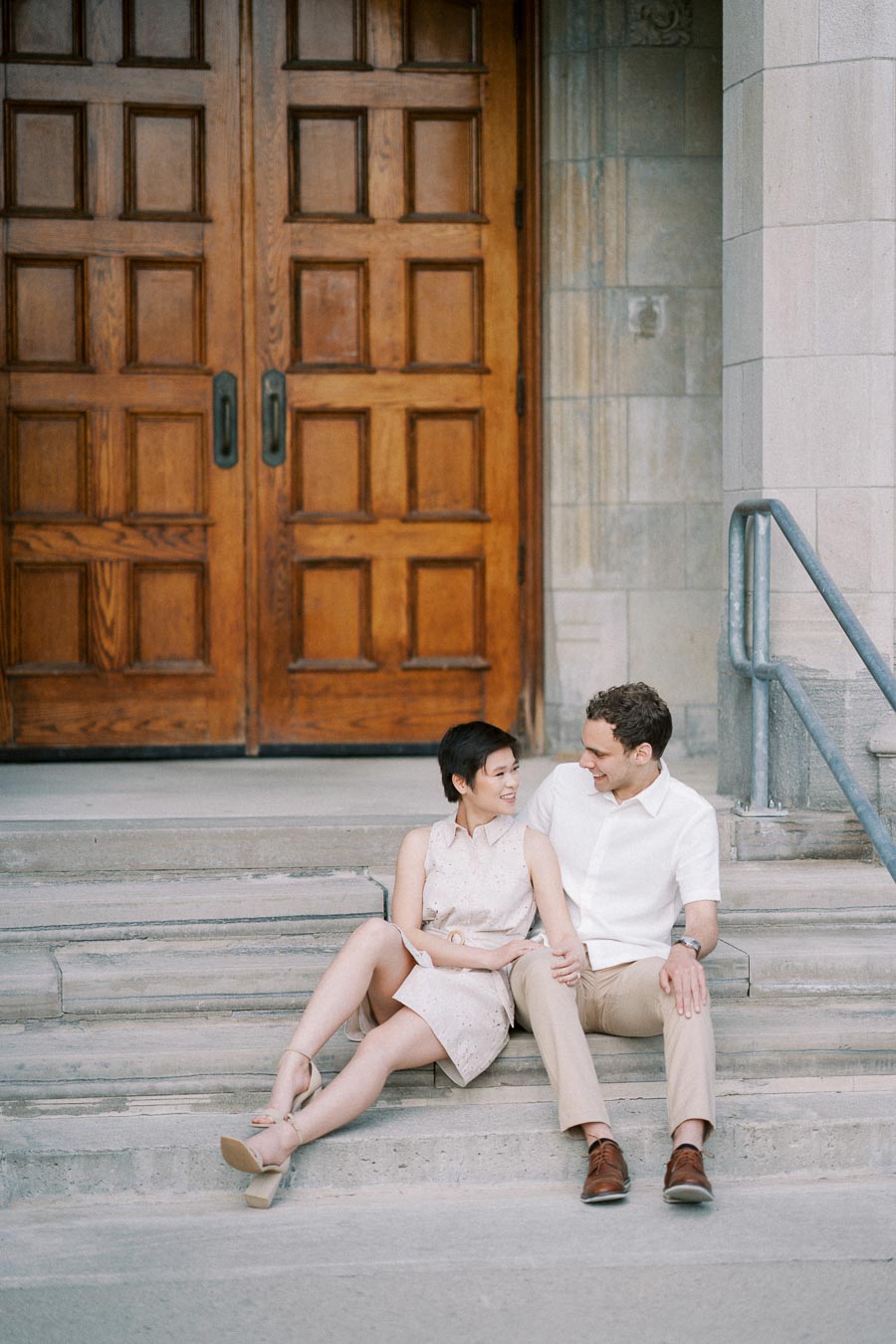 Couple sitting on stone steps in front of large wooden doors, sharing a warm moment with smiles, both dressed in light summer attire.