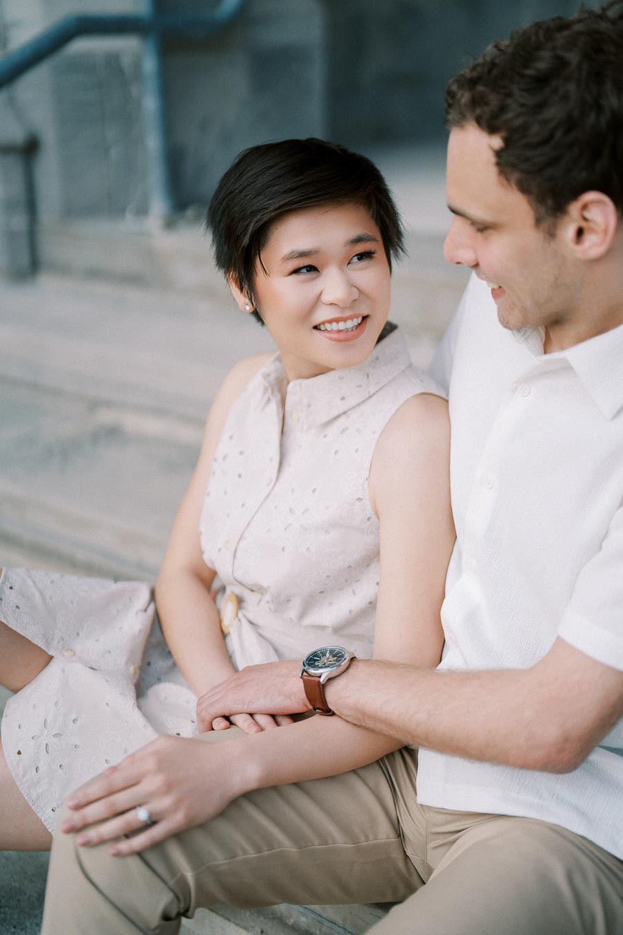 A couple sitting on stone steps, sharing a joyful moment. The woman is wearing a sleeveless dress and the man is in a white shirt, looking at each other and smiling warmly.