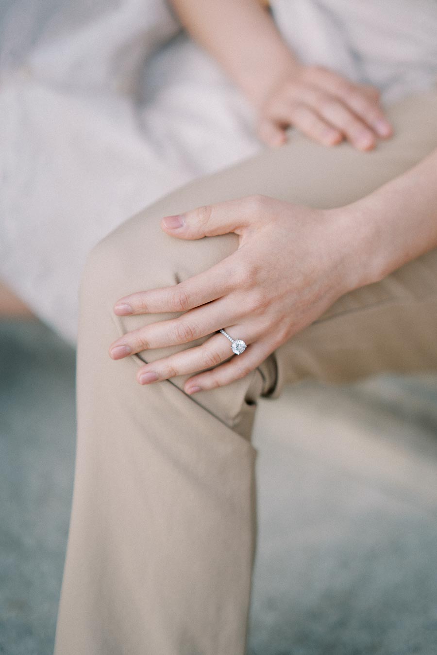 A close-up of a person's hand resting on their knee, showcasing an elegant diamond engagement ring with a round cut stone.