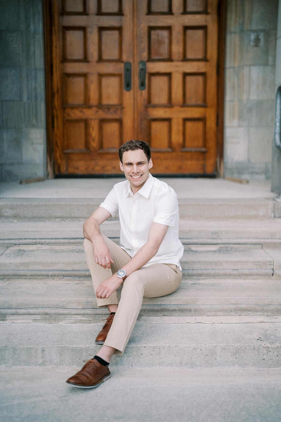 Man sitting on stone steps in front of large wooden doors, wearing a white shirt and beige pants, smiling at the camera.