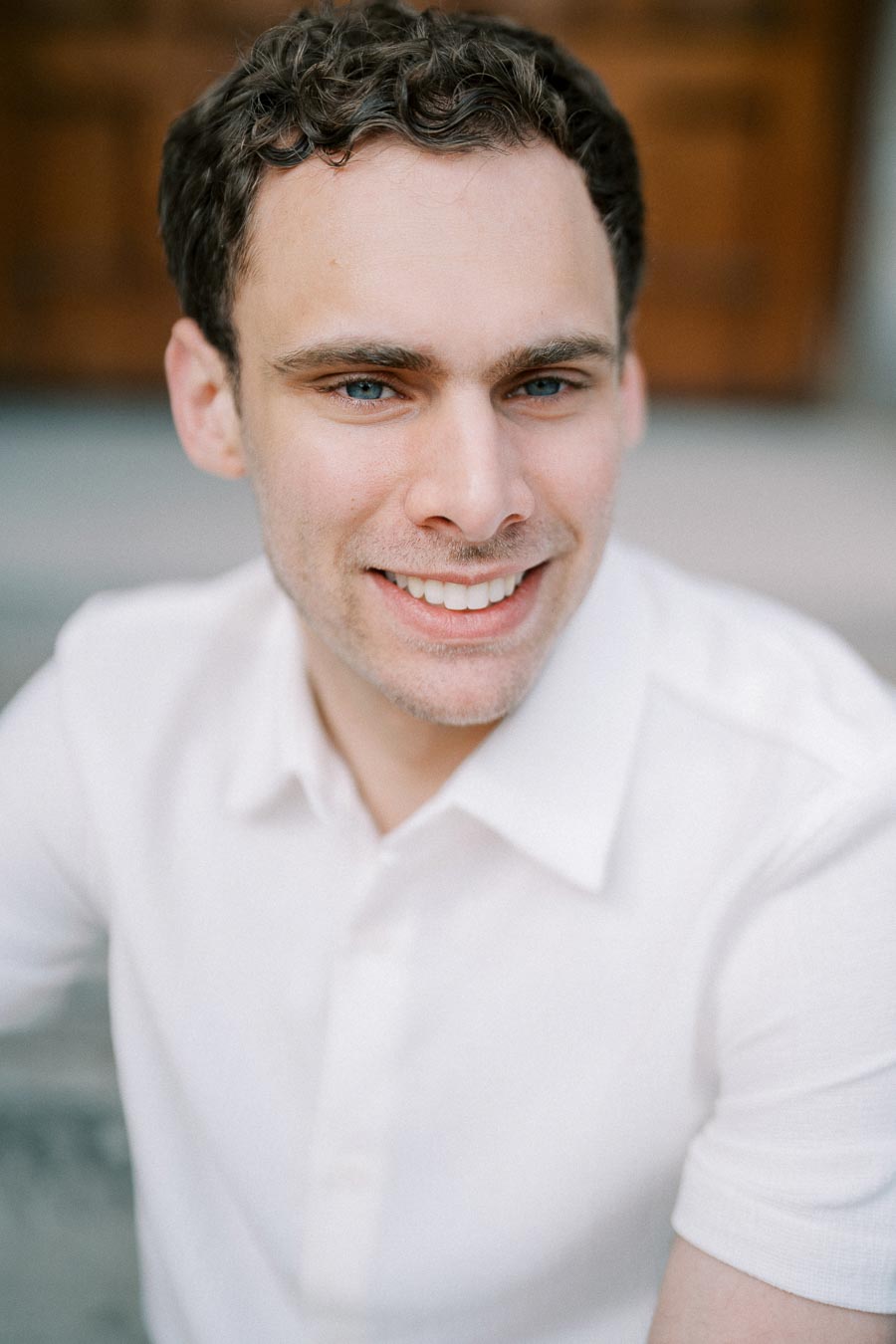 A person smiling while wearing a white shirt, outdoors with a soft focus background.