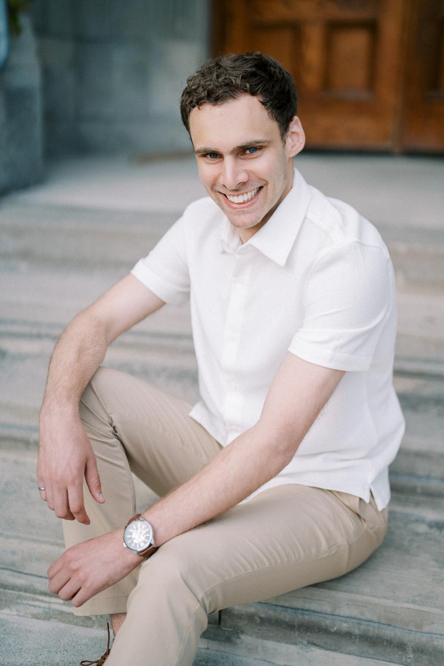 Smiling man sitting on steps wearing a white shirt and beige pants, showcasing a stylish watch.