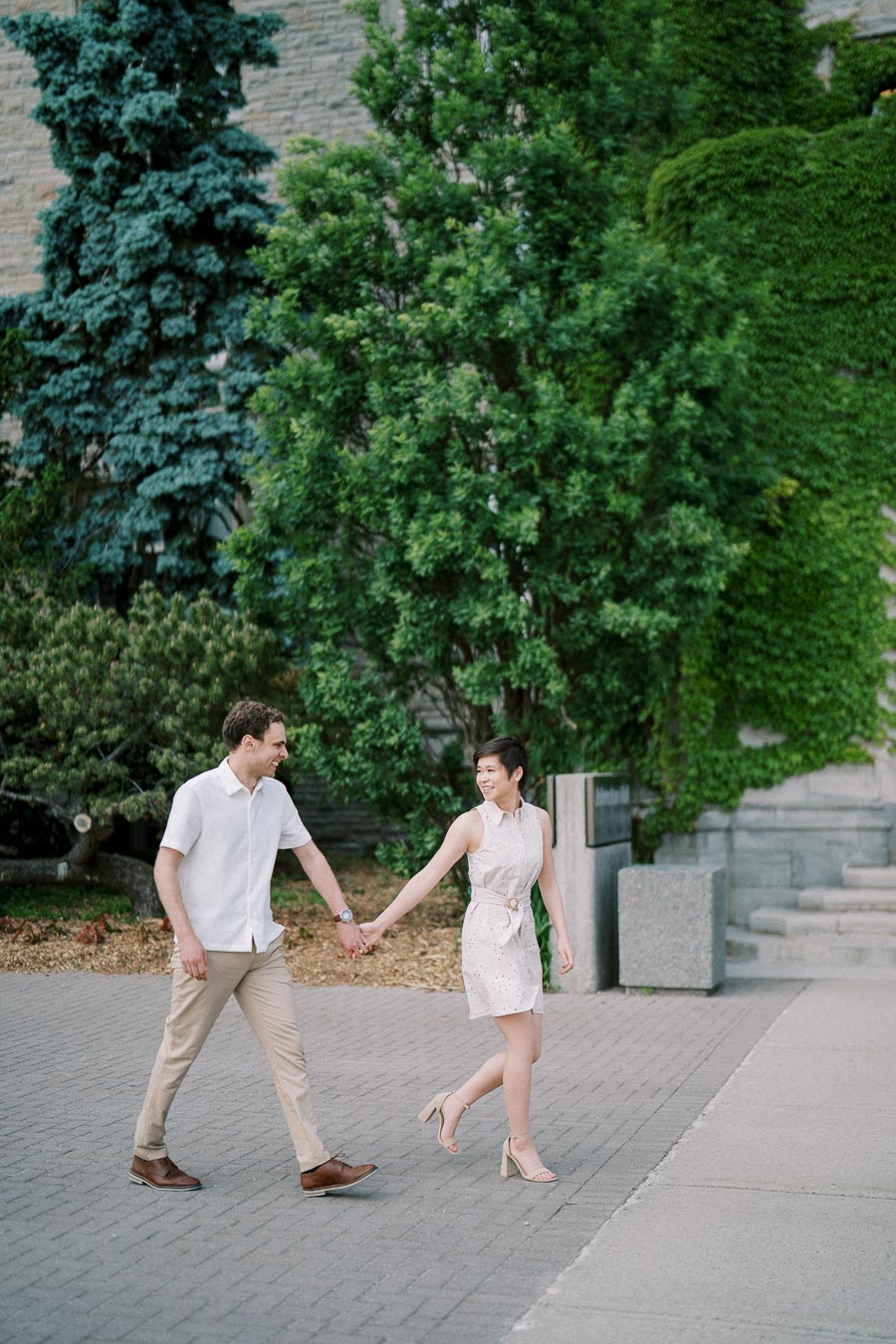A couple holding hands and walking on a paved path surrounded by lush greenery and trees, with a stone wall in the background, conveying a romantic and joyful outdoor scene.