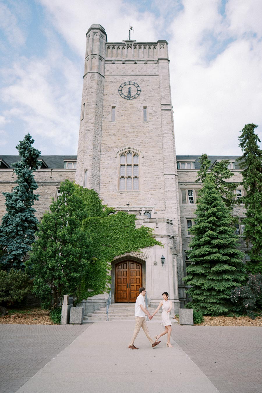 A couple holding hands in front of a historic stone building with a tall clock tower and ivy-covered entrance, surrounded by lush greenery and trees under a partially cloudy sky.