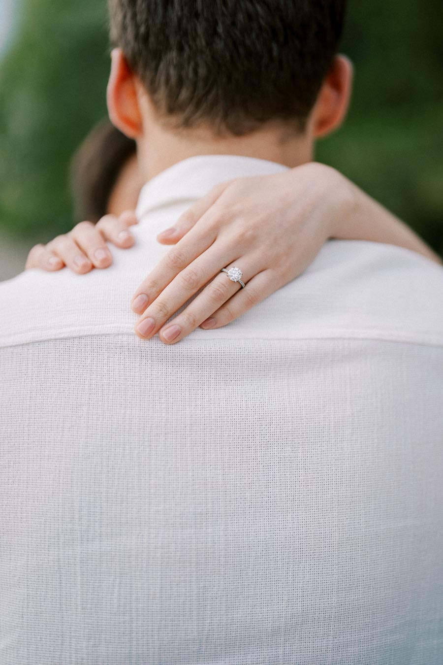 A close-up image of a woman's hand with an engagement ring gently placed on the shoulder of a man wearing a white shirt, symbolizing love and commitment.