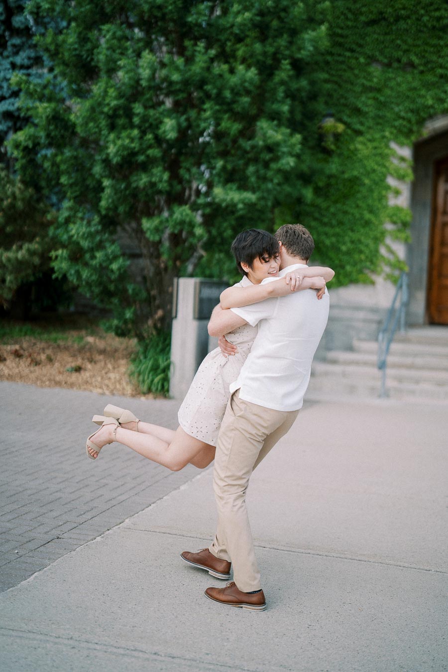 A couple joyfully embraces, with the man lifting the woman off the ground in an outdoor setting. The background features greenery and a building with a wooden door and steps. The woman is wearing a light dress and heels, while the man is dressed in a white shirt and beige pants.
