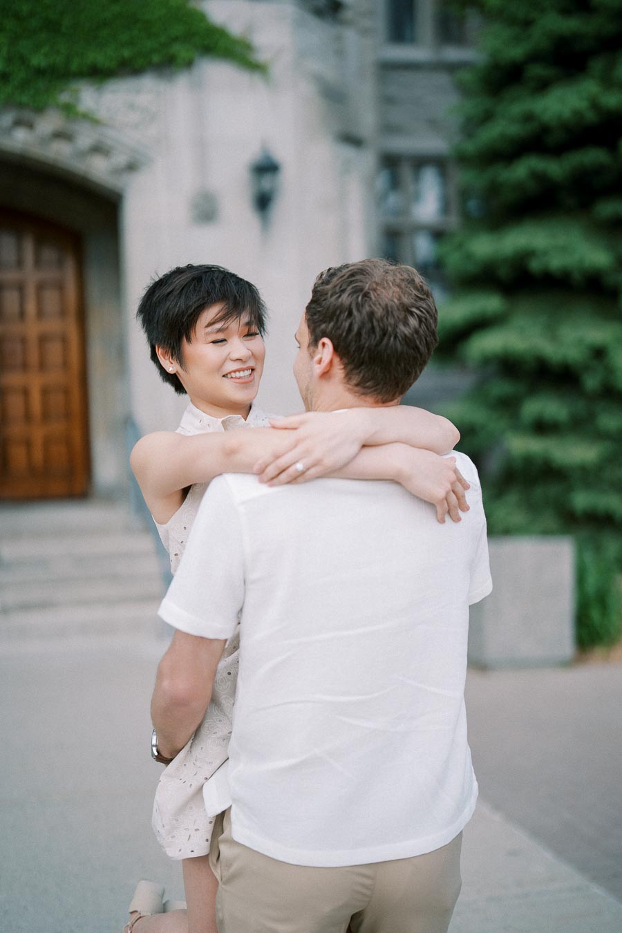 A couple embracing and smiling outside a stone building with greenery and a wooden door in the background.