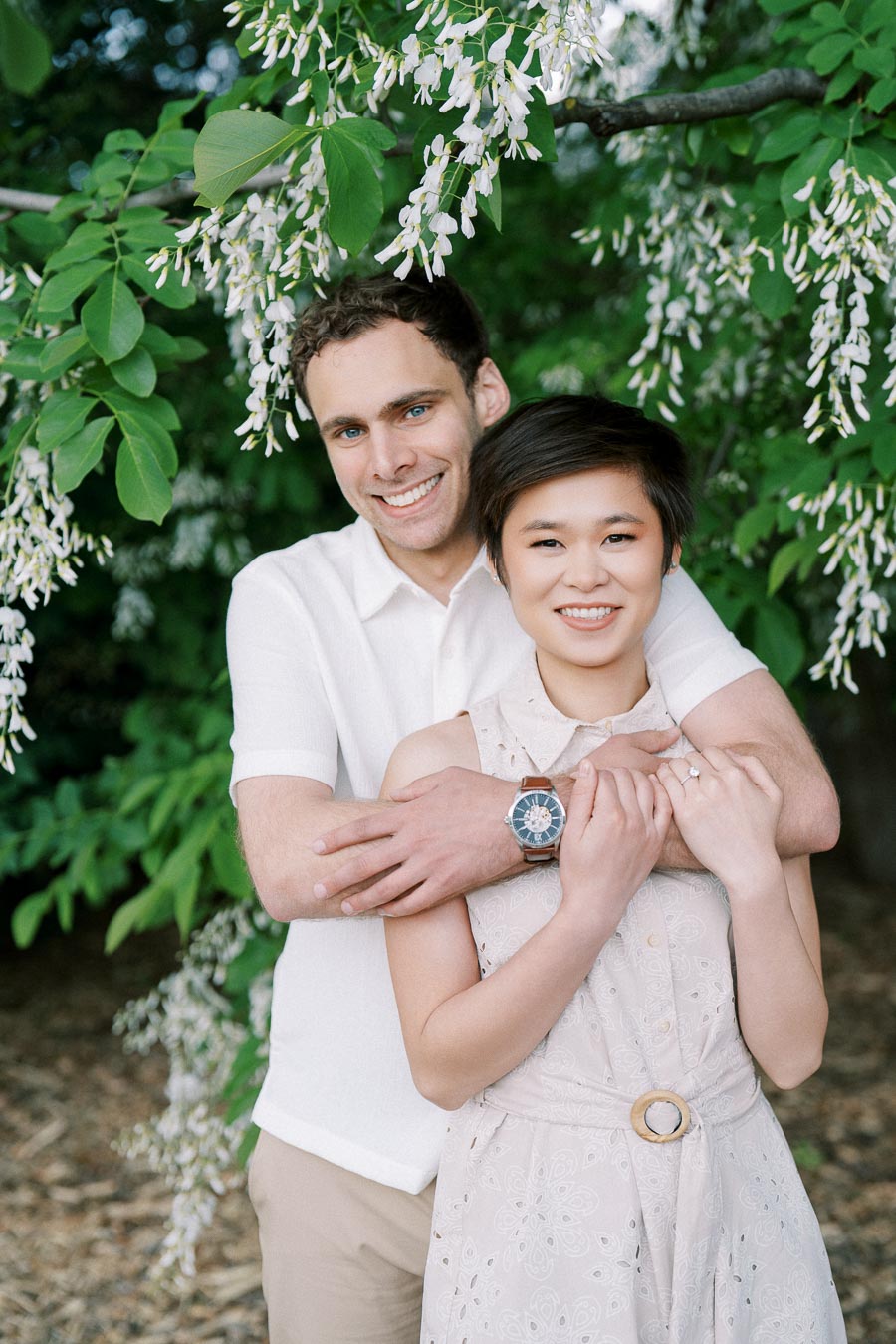 A smiling couple embraces under a blooming tree with green leaves and white flowers, creating a romantic outdoor scene.