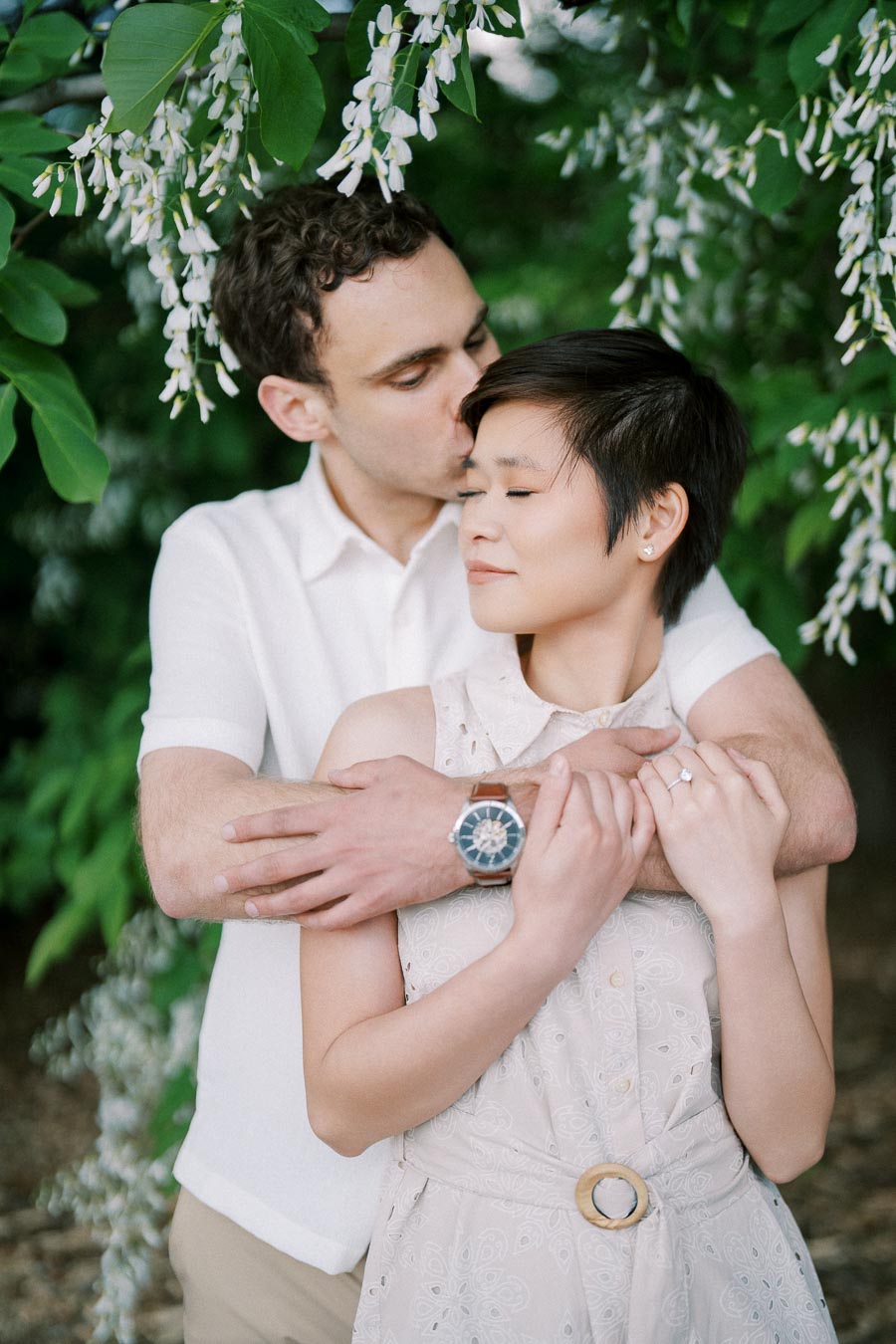 A romantic couple embraces tenderly under a lush canopy of greenery and white blossoms, showcasing love and affection in a serene outdoor setting.