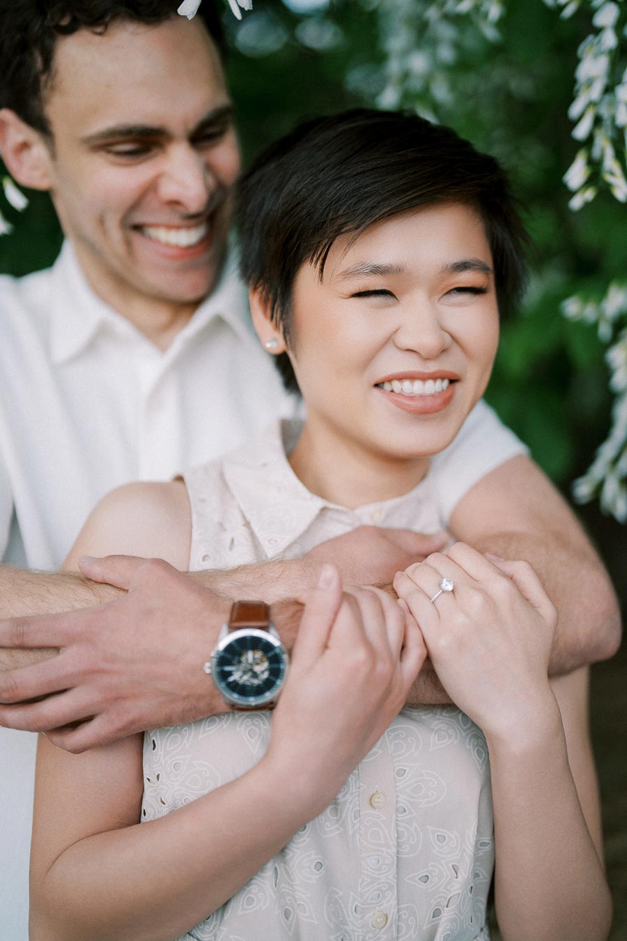 Young couple embracing and smiling under blooming trees, highlighting engagement jewelry and mutual affection.