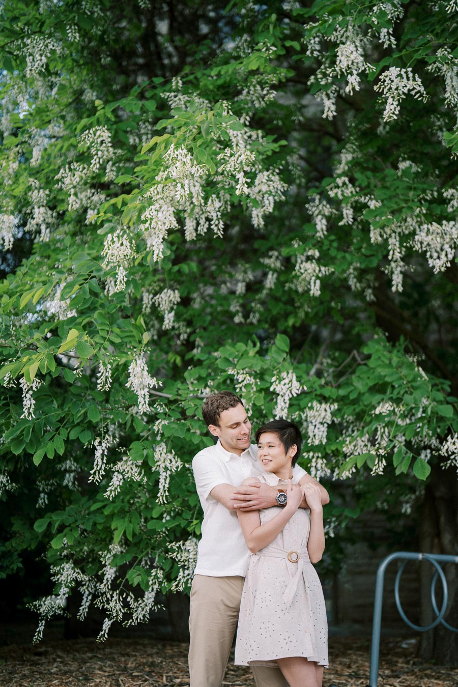 Couple embracing under a lush tree with cascading white flowers, enjoying a serene moment outdoors.