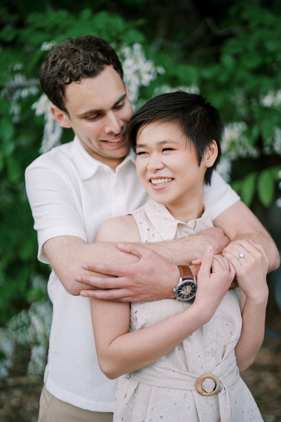 A happy couple embracing in a lush green garden setting, with the man wearing a white shirt and watch, and the woman in a sleeveless dress showing a diamond engagement ring, both smiling joyfully.