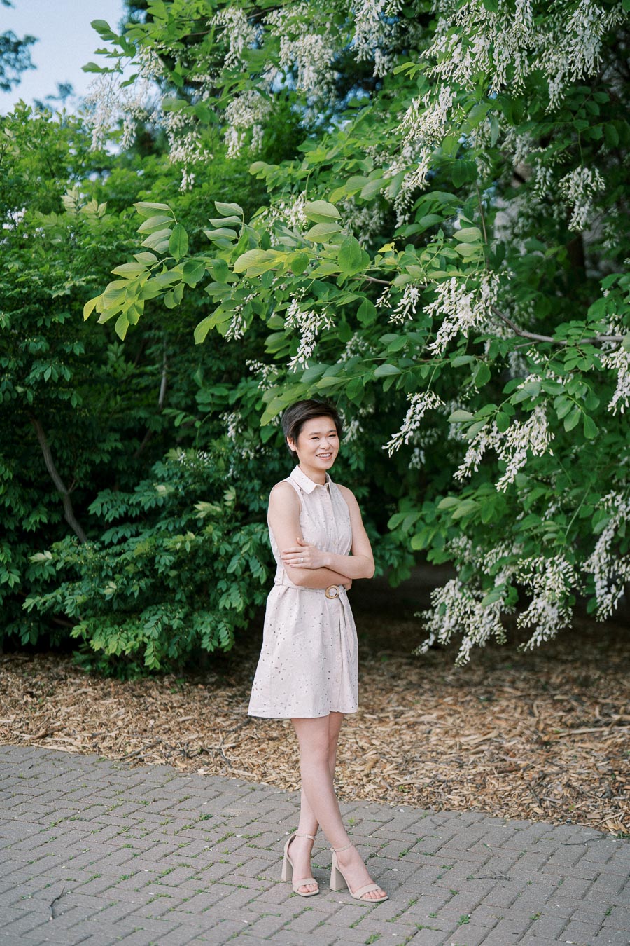 A woman in a light pink dress stands with crossed arms and smiles in a lush garden setting, surrounded by green foliage and blooming white flowers.