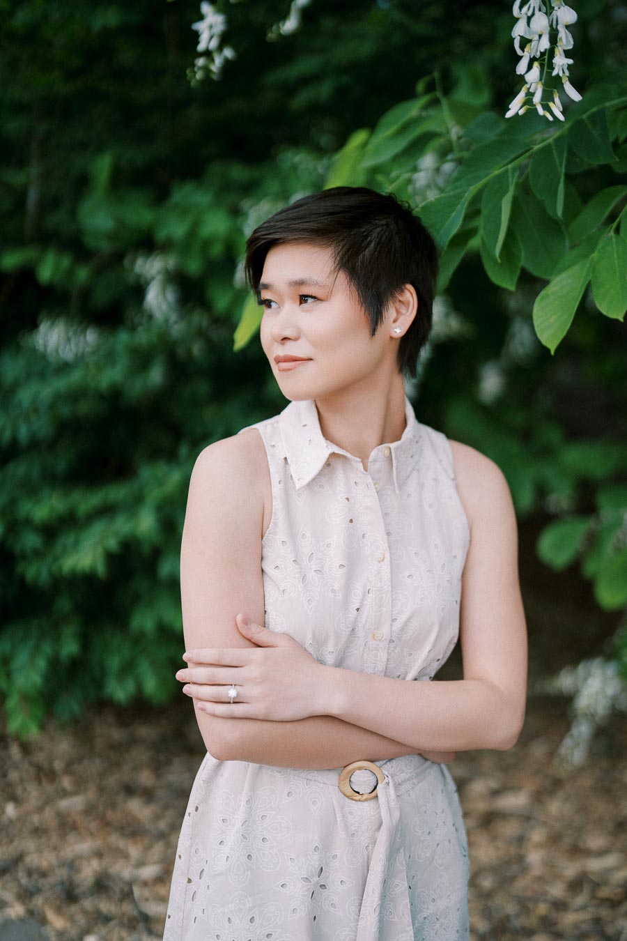 A woman in a sleeveless white dress standing outdoors, surrounded by lush green leaves and plants, with a thoughtful expression.