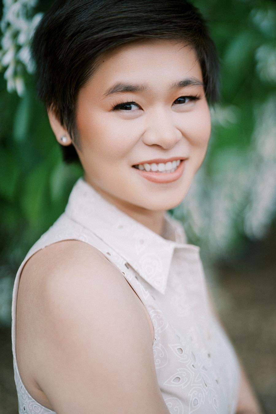 A person smiling outdoors with a blurred green background, wearing a sleeveless, light-colored blouse.