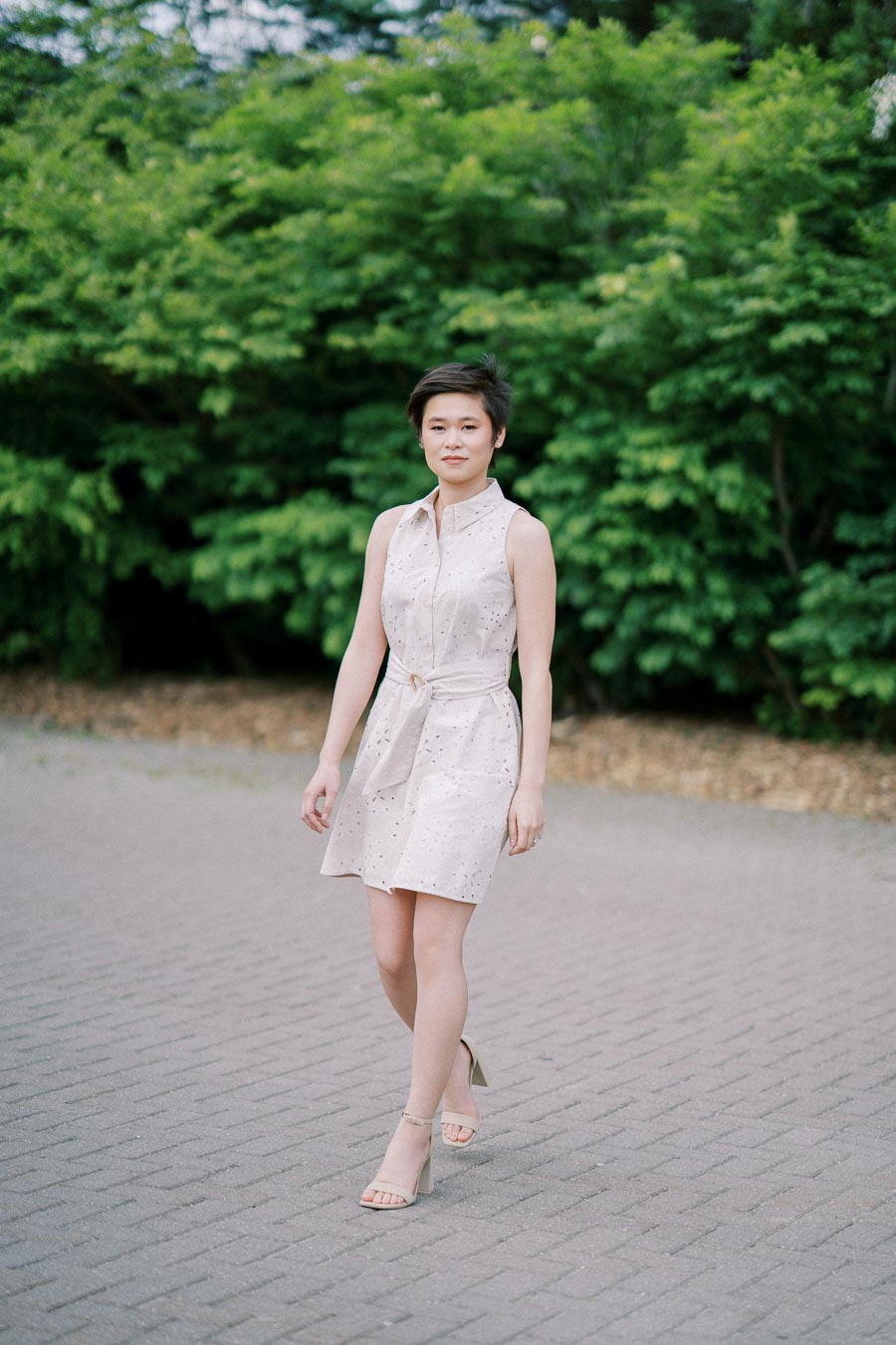 Person in a sleeveless beige dress and sandals walking on a paved path with lush green foliage in the background, capturing a relaxed outdoor moment.