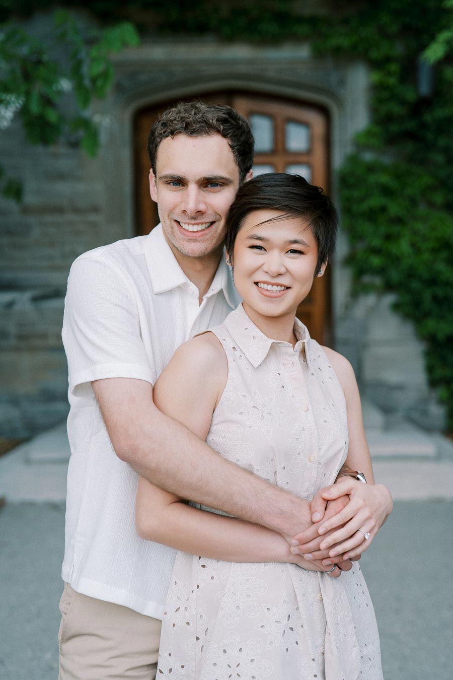 A happy couple wearing light-colored clothes, embracing and smiling in front of a stone building with a wooden door and ivy on the walls.