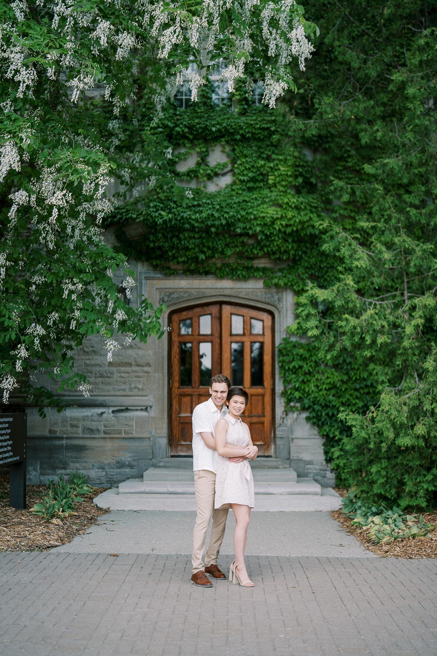 A couple embraces in front of a historic building covered in lush green ivy, featuring a large wooden door. The scene is framed by blooming white flowers and vibrant greenery, creating a romantic and serene atmosphere.