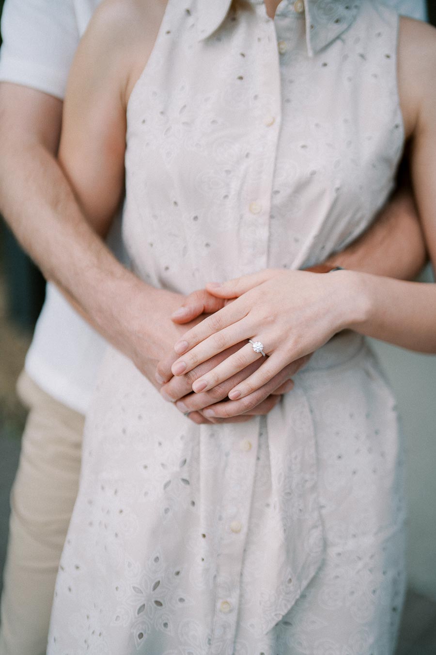A couple embracing warmly, with the woman's hands showing an elegant engagement ring against a light embroidered dress, capturing a moment of love and commitment.