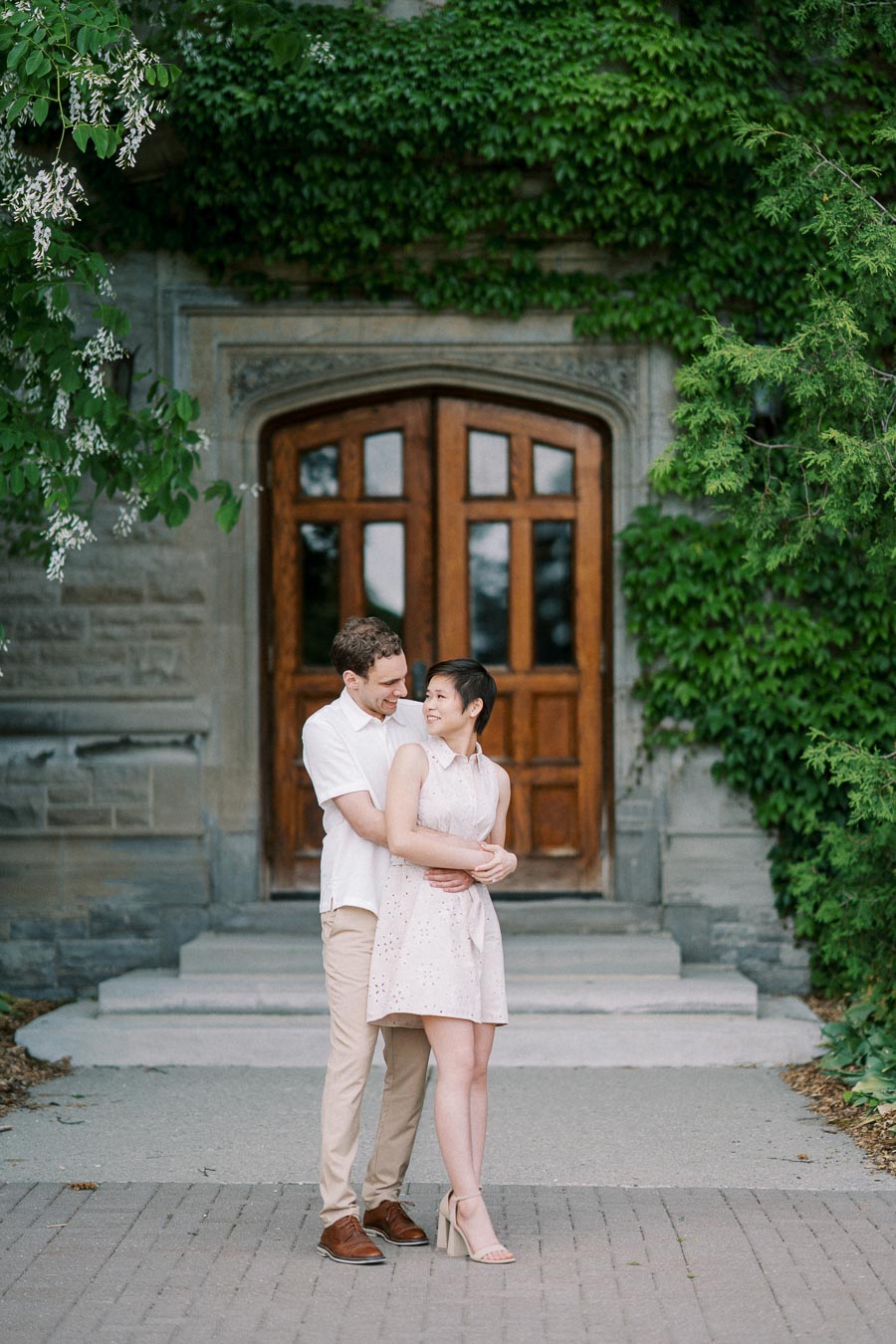 A couple embracing in front of a historic ivy-covered building with a wooden arched doorway, showcasing a romantic and serene outdoor setting.