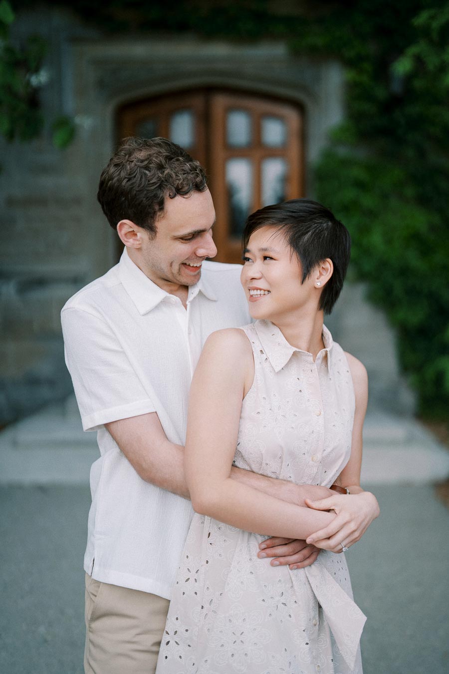 A happy couple embracing outdoors in front of a rustic stone building, with greenery in the background.