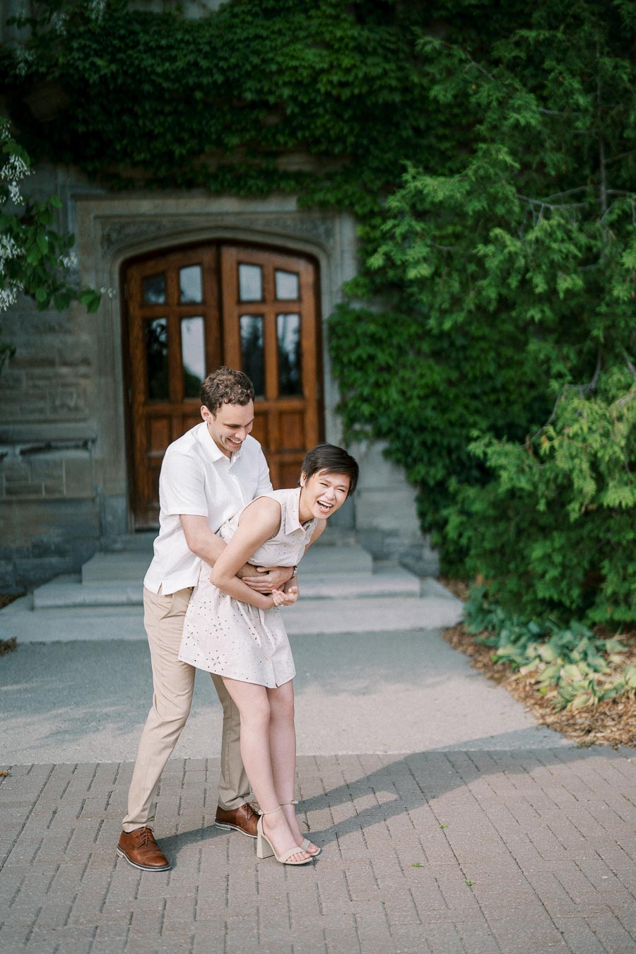 A couple joyfully embracing in a garden setting with lush greenery and a wooden door in the background, the woman in a white dress and the man in a white shirt and beige pants, both laughing and enjoying a playful moment on a sunlit pathway.