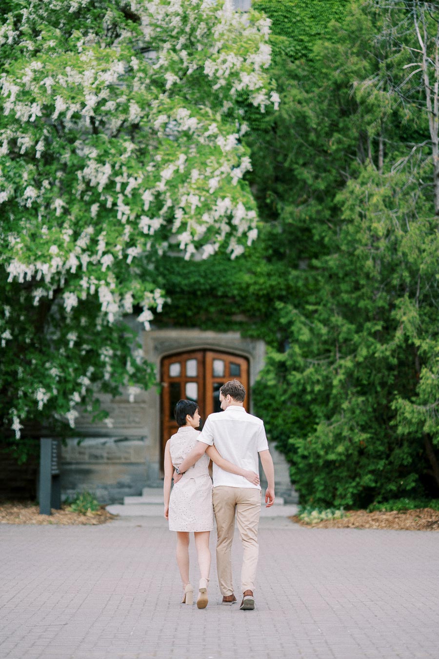 A couple walking arm in arm towards a wooden door surrounded by lush greenery and blooming trees in a serene outdoor setting.