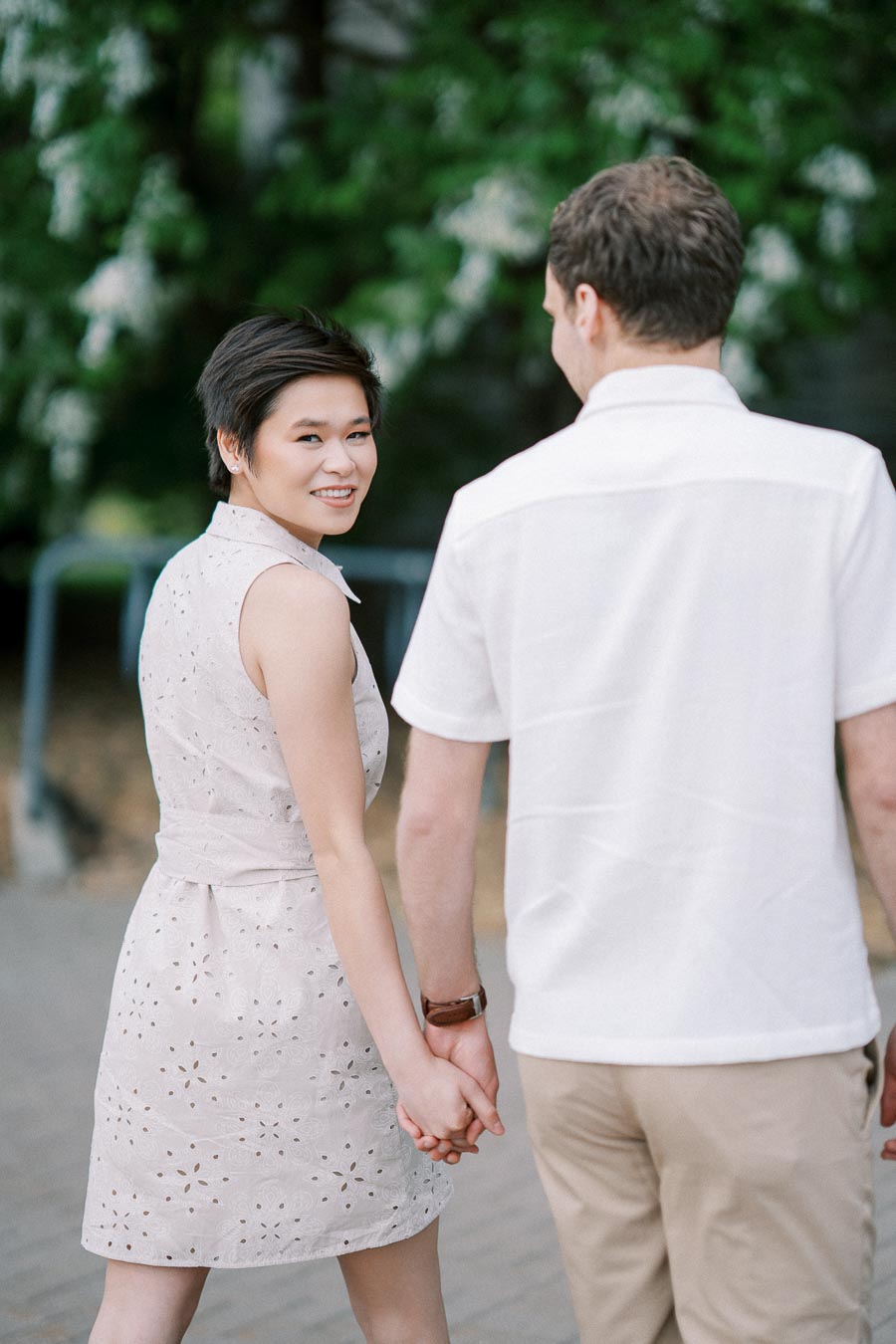 A couple holding hands and walking outside on a sunny day, with green foliage in the background. The woman is smiling and wearing a light-colored summer dress, while the man wears a white shirt and beige pants.