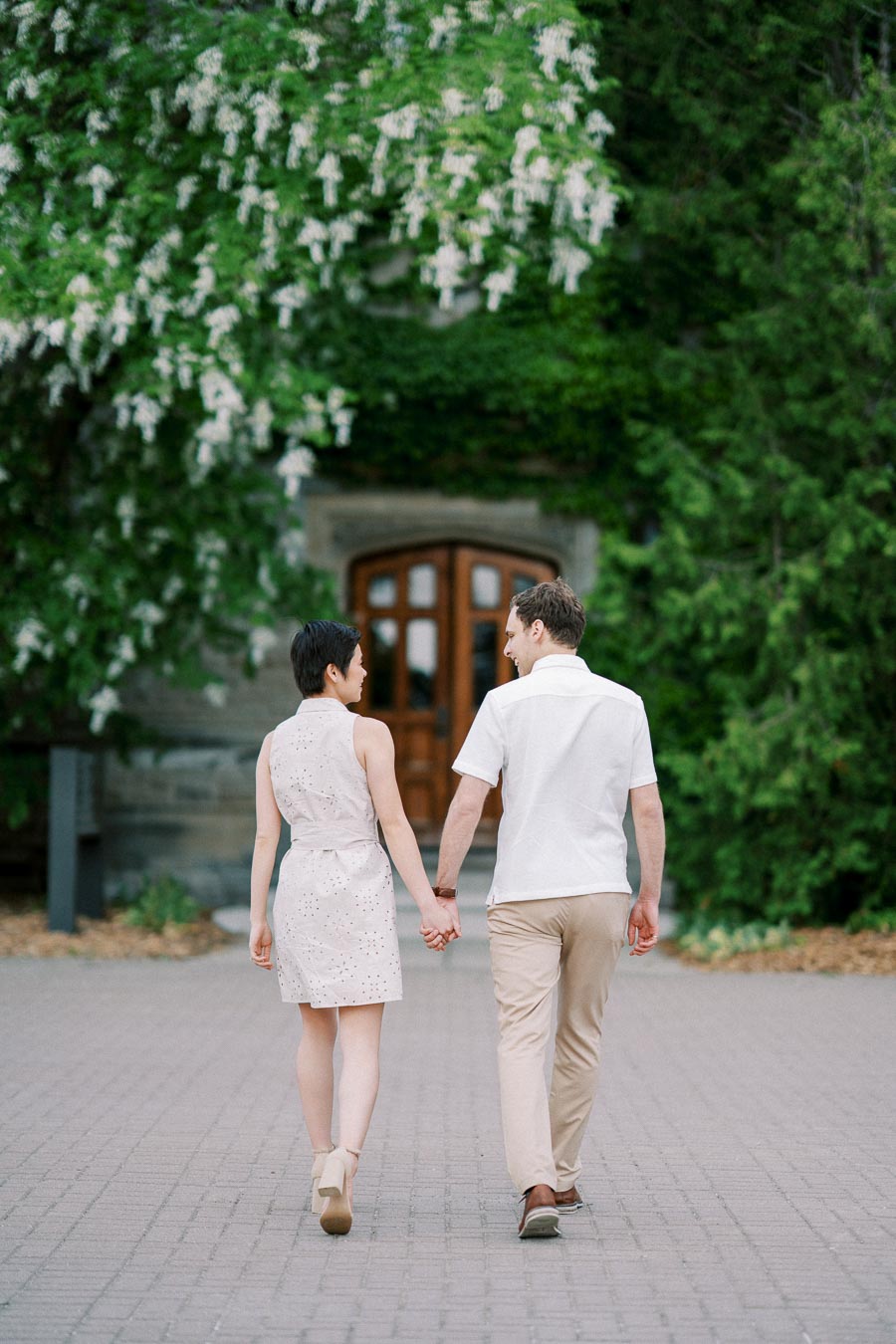 A couple holding hands and walking towards a wooden door, surrounded by lush green foliage and white flowering trees.