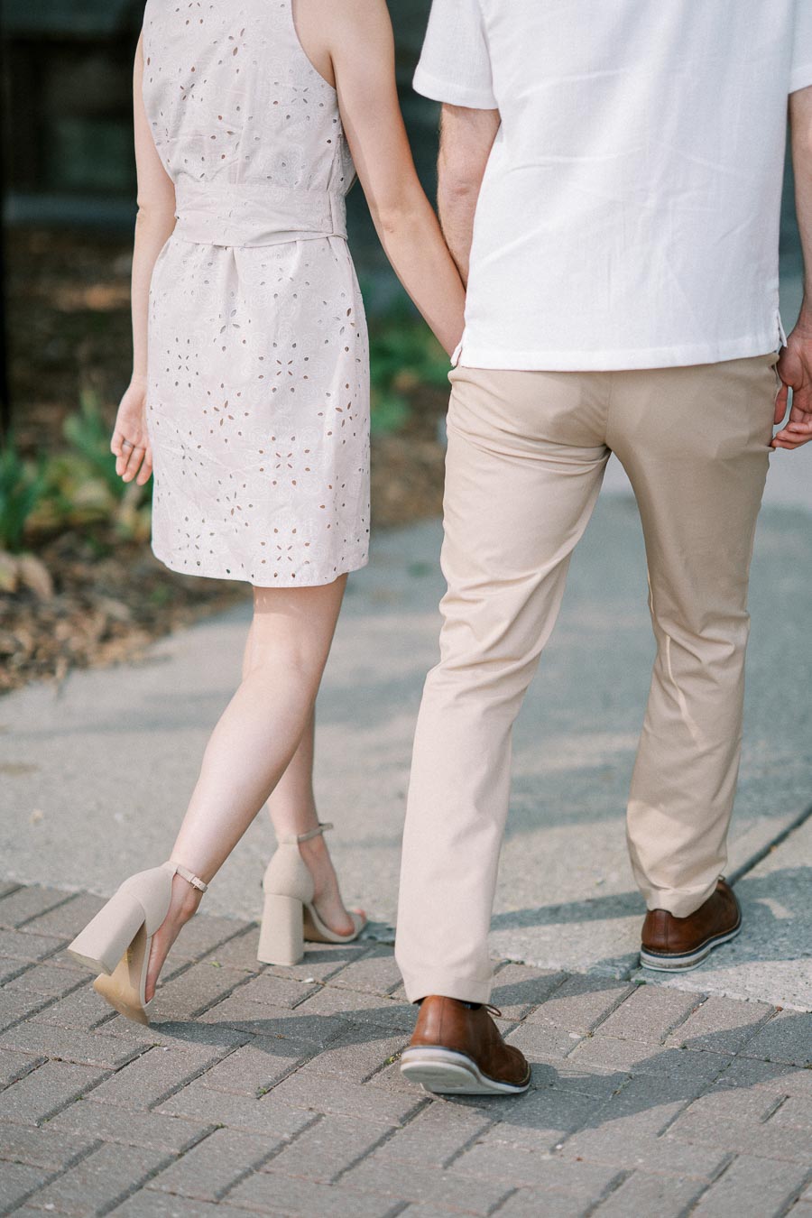 Rear view of a couple holding hands while walking on a paved path, with the woman wearing a white eyelet dress and beige heels, and the man in beige pants and brown shoes.