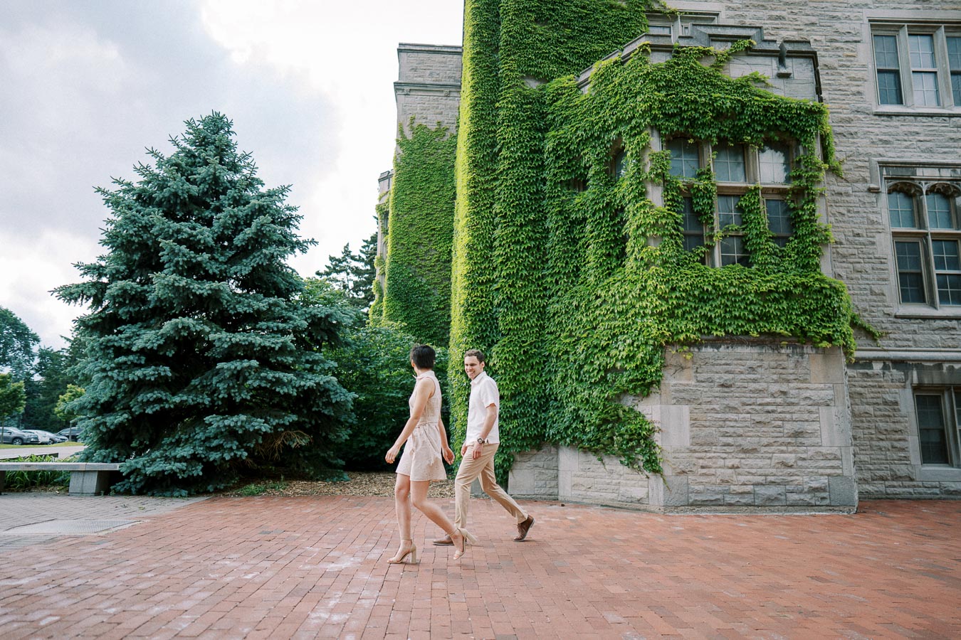 A couple walking on a brick pathway in front of an ivy-covered historic building with stone architecture.