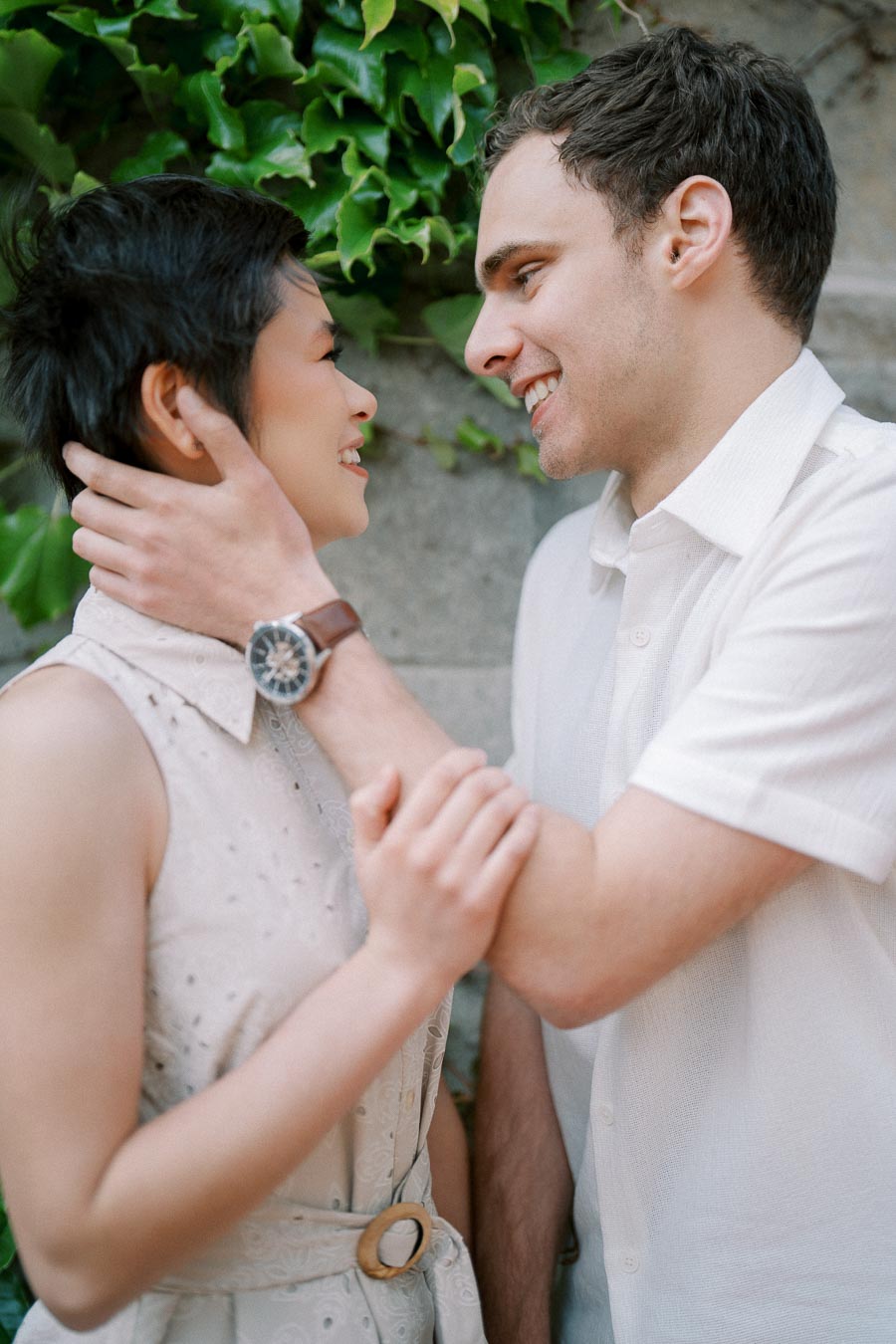 A happy couple smiling and looking at each other, standing against a leafy backdrop.