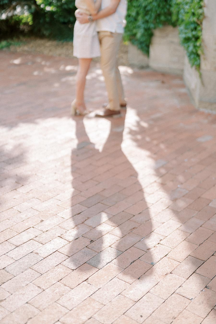 A couple embracing on a sunlit brick path with their shadow cast on the ground, surrounded by lush greenery.