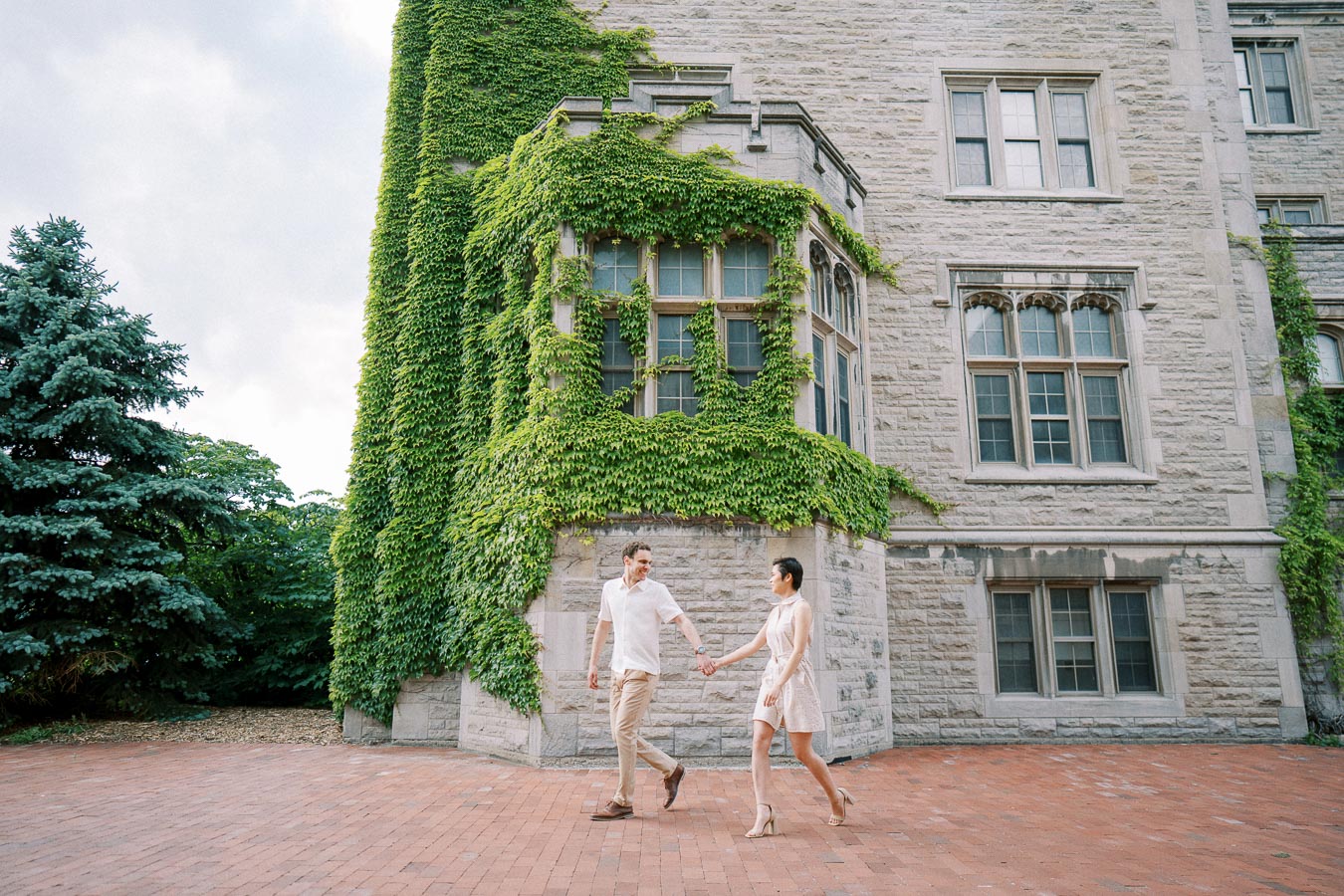 A couple walking hand in hand on a brick pathway in front of an ivy-covered stone building, exuding a sense of romance and elegance.