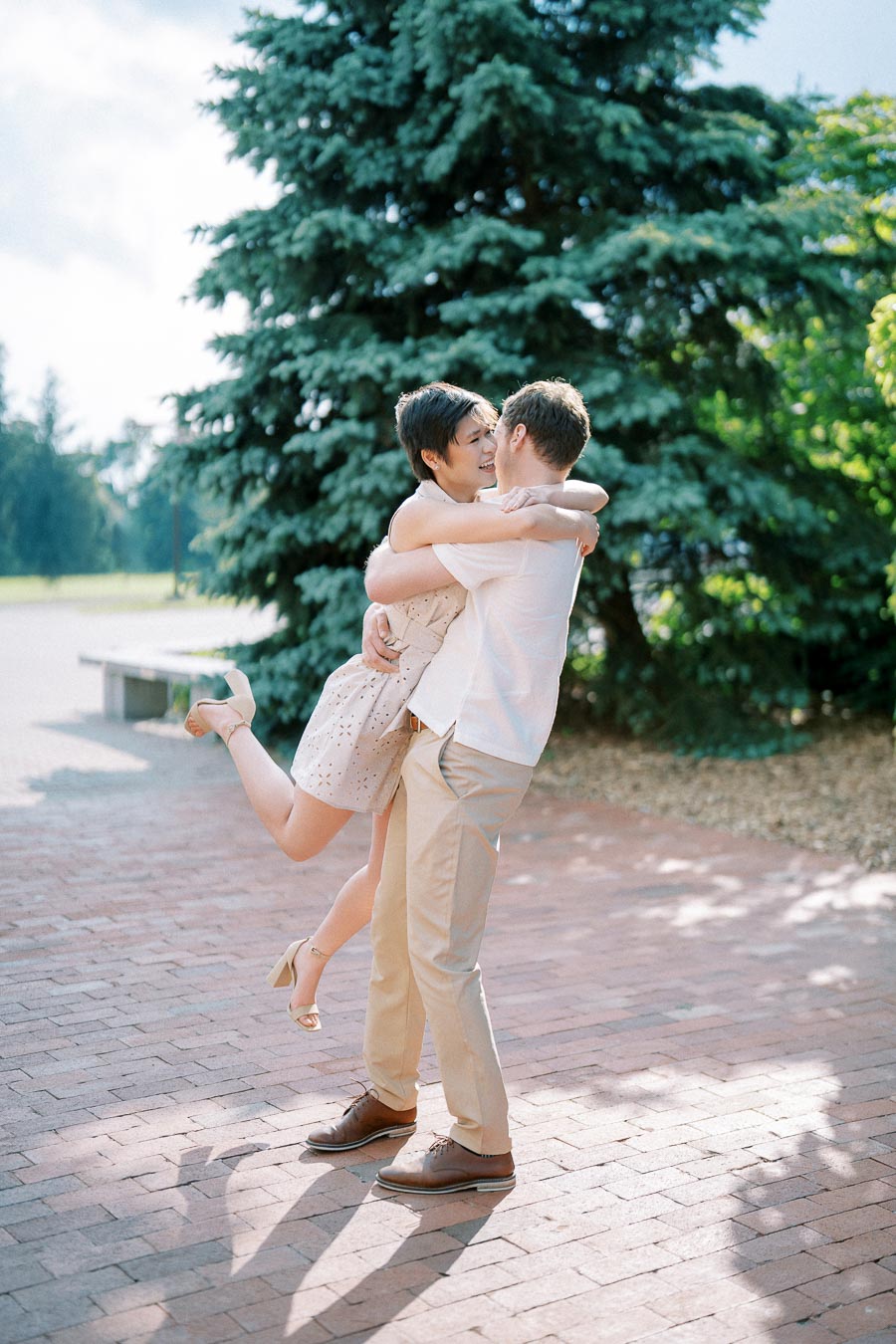 A joyful couple embracing on a sunny day, surrounded by lush greenery, with the man lifting the woman off the ground on a brick pathway.