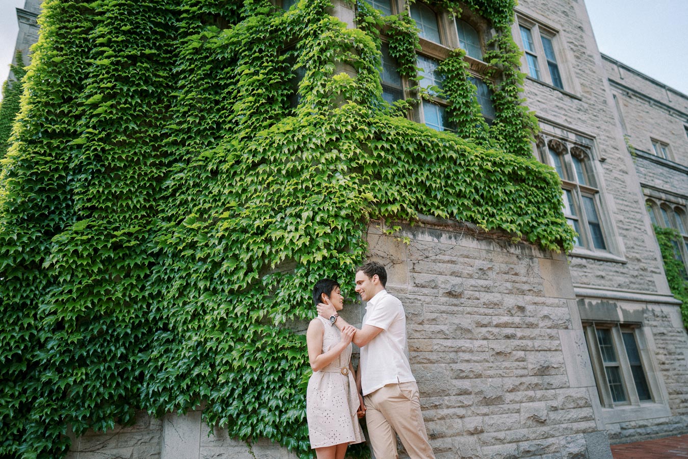 A couple standing closely in front of a historic stone building covered with lush green ivy.