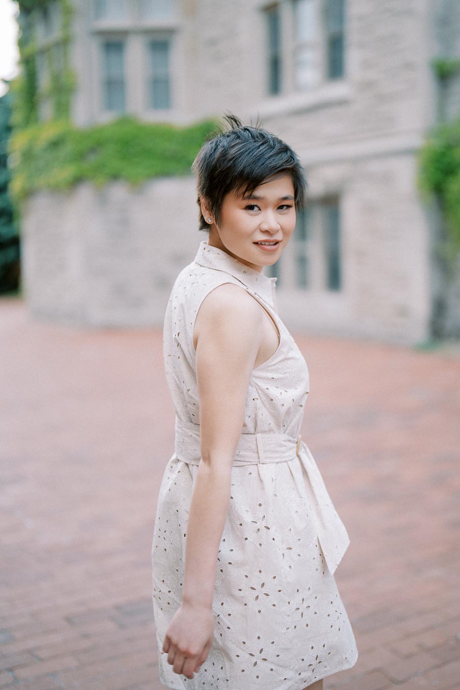 A woman in a white sleeveless dress poses outside a historic building with ivy-covered walls.