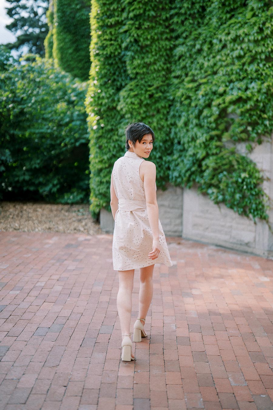 Young woman in a light dress walking on a brick path, surrounded by lush green ivy-covered walls.