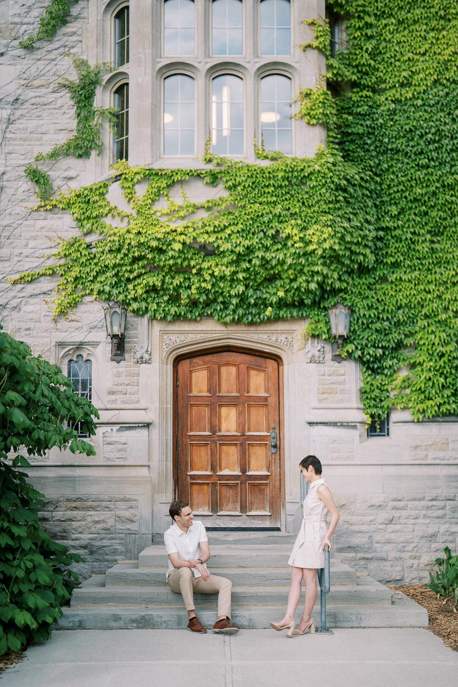A man and woman engaged in conversation on the steps of a historic ivy-covered stone building with a wooden arched door and tall windows.