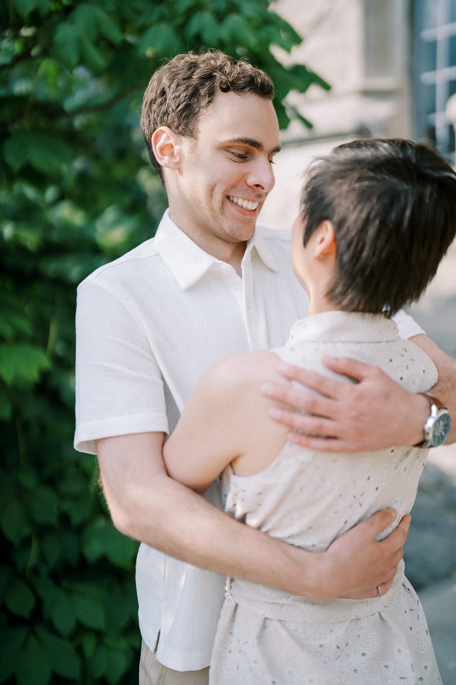 A joyful couple embracing outdoors with lush green foliage in the background, both wearing light, summery clothing.